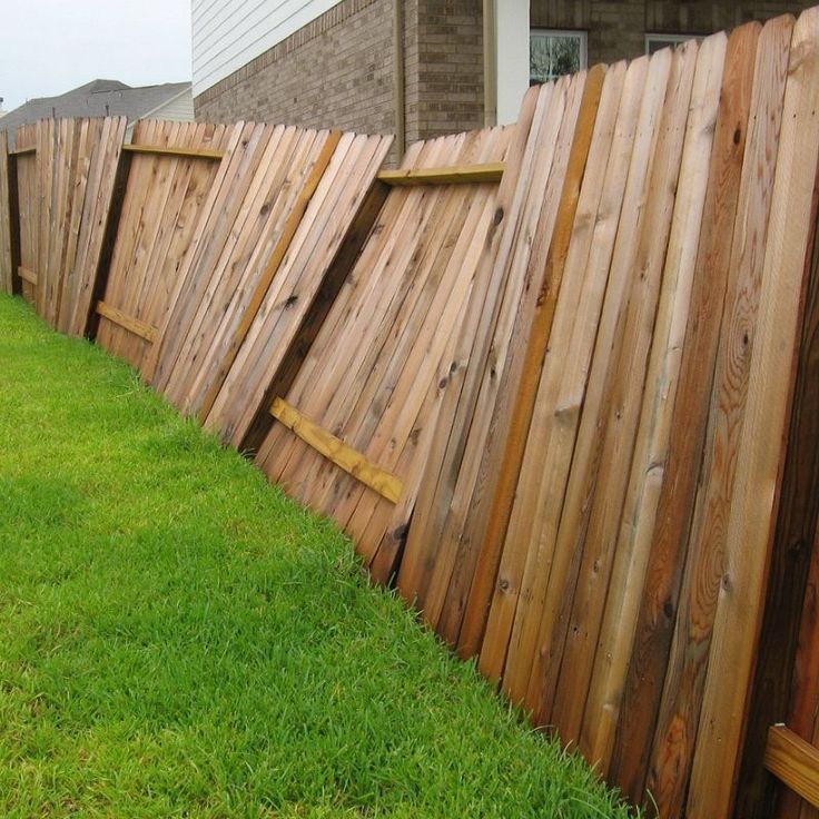 Wooden fence panels are leaning and partially detached, tilting onto a grassy lawn beside a brick house.