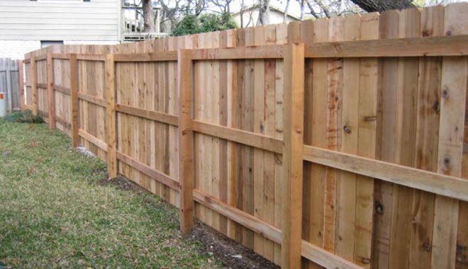 A wooden privacy fence runs along a grassy backyard with vertical planks and horizontal support beams.