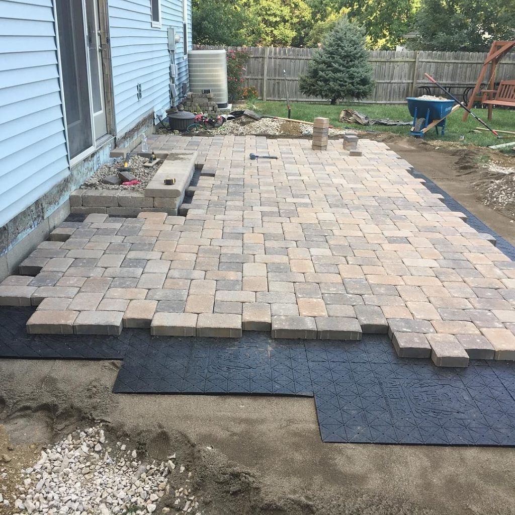 Patio construction in progress with interlocking pavers being installed beside a house, tools and materials visible nearby.