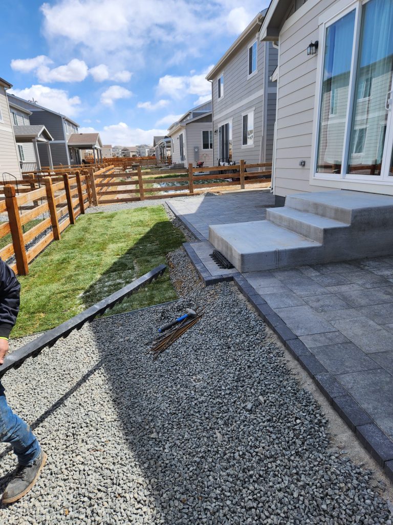 Backyard under construction with gravel, pavers, and concrete steps near a lawn and wooden fence on a sunny day.
